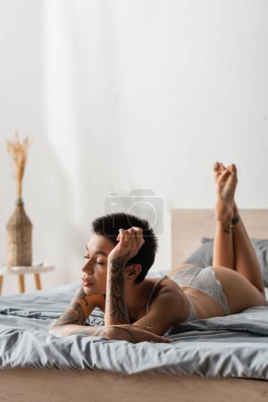young appealing woman with sexy tattooed body and short brunette hair laying on grey bedding in lingerie near bedside table and wicker vase with spikelets on blurred background in bedroom