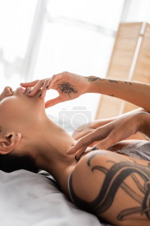 Foto de Vista parcial de la mujer joven y apasionada tocando los labios y el cuerpo tatuado sexy, mientras que la colocación y posando en lencería sobre ropa de cama gris en el dormitorio moderno, fotografía boudoir - Imagen libre de derechos