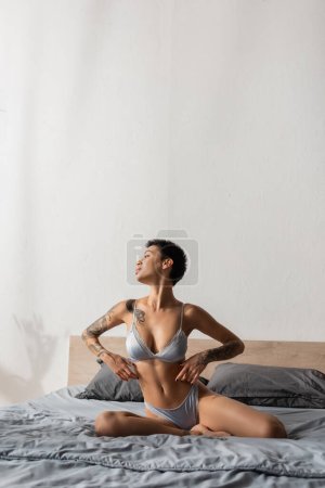 irrésistible femme en lingerie de soie, avec cheveux bruns courts et corps tatoué sexy regardant loin tout en posant sur la literie grise près des oreillers dans la chambre moderne, photographie boudoir