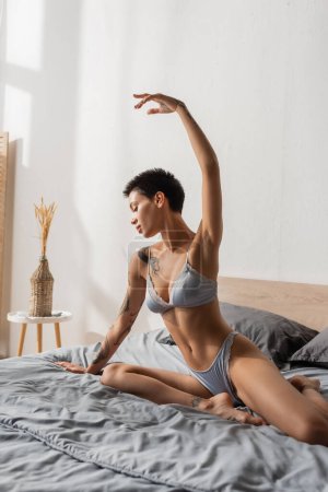 stunning tattooed woman with sexy slender body and short brunette hair posing with raised hand on grey bedding near pillows, bedside table and vase with spikelets in bedroom, boudoir photography