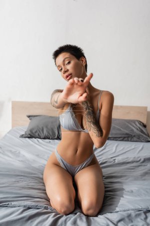 young and intriguing woman in lingerie, with short brunette hair and sexy tattooed body looking at camera and showing stop gesture on grey bedding near pillows in bedroom, blurred foreground