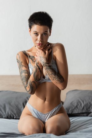 seductive and intriguing woman with sexy tattooed body and short brunette hair looking at camera while posing in silk lingerie on grey bedding near pillows on blurred background, boudoir photography
