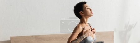 Photo for Side view of captivating and sexy woman with tattooed body and short brunette hair touching straps of grey silk bra while looking away near white wall in modern bedroom, banner - Royalty Free Image