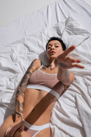 Photo for Top view of intriguing young woman in beige lingerie, with short brunette hair and sexy tattooed body laying on white bedding with outstretched hand and looking at camera in studio - Royalty Free Image