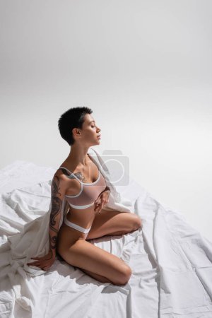 vista de ángulo alto de mujer deseable y joven en lencería beige, con pelo corto morena y cuerpo tatuado sentado en pose seductora sobre ropa de cama blanca sobre fondo gris, fotografía erótica