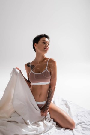 mujer joven, tatuada y apasionada en lencería beige, con cuerpo sexy y pelo corto morena sosteniendo sábana blanca y mirando hacia otro lado en el estudio sobre fondo gris, fotografía erótica