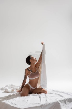 mujer delgada e impresionante en lencería beige, con pelo corto morena y cuerpo tatuado sexy sosteniendo sábana en la mano levantada mientras está sentado sobre ropa de cama blanca y fondo gris, arte de la seducción