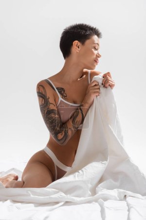 junge, sinnliche und tätowierte Frau mit sexy Körper und kurzen brünetten Haaren sitzt in beiger Dessous und hält weißes Bettlaken auf grauem Hintergrund im Studio, erotische Fotografie