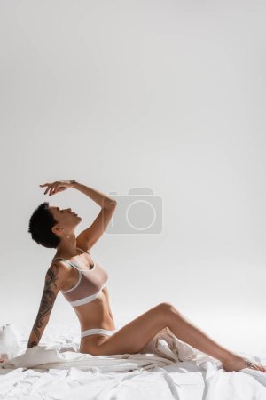 Téléchargez les photos : Vue latérale de jeune femme sexy et charmante en lingerie beige, avec de courts cheveux bruns et un corps tatoué assis avec la main au-dessus de la tête sur une literie blanche et un fond gris, photographie érotique - en image libre de droit