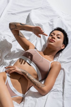 vista de ángulo alto de mujer joven y apasionada en lencería beige, con cuerpo tatuado sexy y pelo corto morena tendido en ropa de cama blanca y mirando a la cámara en el estudio, fotografía erótica