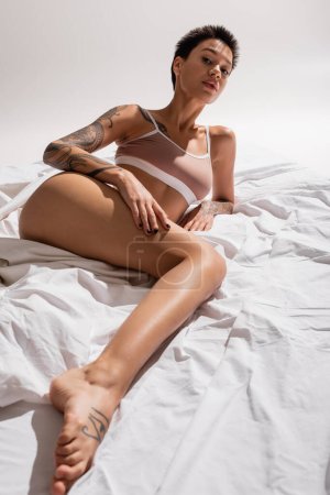 young intriguing and tattooed woman in beige bra, with sexy body and short brunette hair looking at camera on grey background, art of seduction, erotic photography