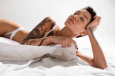 mujer joven y sensual con pelo corto morena y cuerpo tatuado sexy posando en lencería con los ojos cerrados sobre ropa de cama blanca y fondo gris, fotografía erótica, arte de la seducción