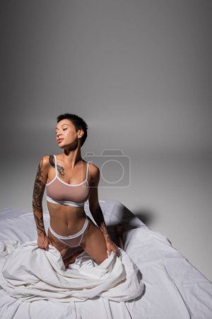 young and provocative woman with short brunette hair and sexy tattooed body, wearing beige lingerie and posing on knees on white bedding and grey background, erotic photography