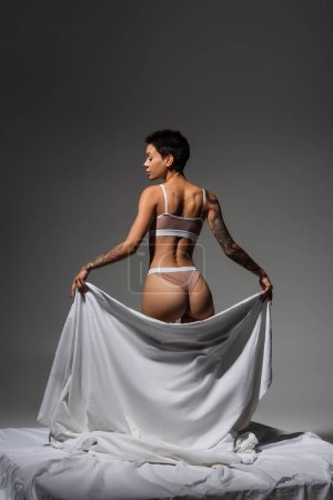 back view of stunning and sexy woman in beige lingerie, with short brunette hair and tattooed body holding white bed sheet while standing on grey background in studio, art of seduction