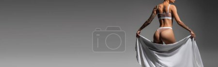 partial view of young and sexy woman with tattooed body and sexy buttocks standing in beige lingerie while posing with white bed sheet on grey background, erotic photography, banner