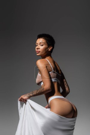 young appealing woman with tattooed body, short brunette hair and sexy buttocks, wearing beige lingerie and looking at camera while posing with white bed sheet on grey background