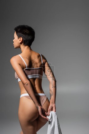 Photo for Young and desirable woman with short brunette hair and sexy tattooed body holding white bed sheet while posing in beige lingerie in studio on grey background, erotic photography - Royalty Free Image