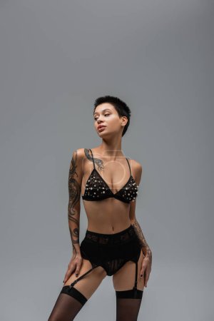 tattooed and expressive woman with short brunette hair and tattooed body posing in bra with pearl beads, lace panties, garter belt and black stockings and looking away on grey background