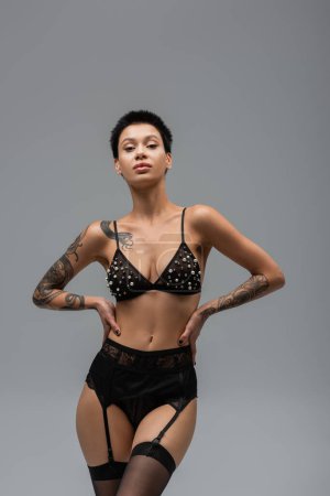 sexy mujer tatuada en lencería glamour negro, sujetador con perlas, bragas de encaje, liguero y medias posando con las manos en las caderas y mirando a la cámara sobre fondo gris