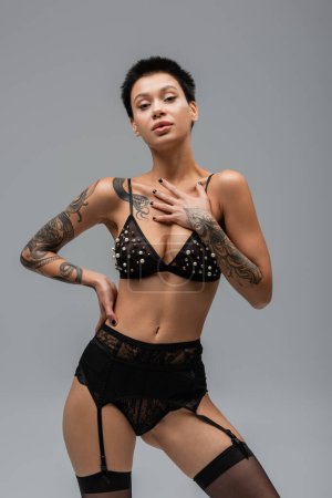 provocative woman with sexy tattooed body and short brunette hair, in bra with pearl beads, black panties, garter belt and stockings posing with hand on hip and looking at camera on grey background