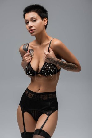 atractiva mujer tatuada con cuerpo tatuado sexy y pelo corto morena posando en bragas de encaje negro, liguero y medias mientras toca correas de sujetador con cuentas de perlas sobre fondo gris