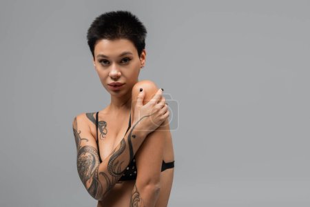 mujer joven y apasionada con pelo corto morena y cuerpo tatuado sexy mirando a la cámara mientras posa en sujetador negro con perlas sobre fondo gris en el estudio, arte de la seducción
