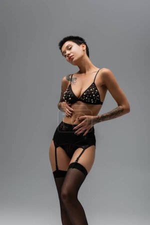 passionate and stylish tattooed woman with short brunette hair posing in bra with pearl beads, black lace panties, garter belt and stocking on grey background in studio, erotic photography