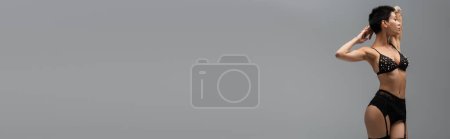 Téléchargez les photos : Pleine longueur de femme tentante et sexy avec corps tatoué posant avec les mains derrière la tête en soutien-gorge avec perles de perles, culotte en dentelle et porte-jarretelles noir sur fond gris, bannière - en image libre de droit