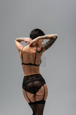 Photo for Back view of irresistible woman with tattooed body and sexy buttocks wearing black bra, lace panties, garter belt and black stockings while standing with hands behind neck on grey background - Royalty Free Image