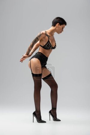 Photo for Full length of sexy young woman with tattooed body posing in bra with pearl beads, lace panties, garter belt, black stockings and high heels on grey background in studio - Royalty Free Image
