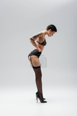 Photo for Full length of stunning and slender woman with sexy tattooed body, wearing bra with pearl beads, lace panties, garter belt and black stockings while posing on high heels on grey background - Royalty Free Image