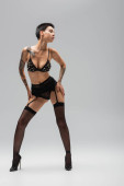 full length of expressive woman with sexy tattooed body wearing black bra with pearl beads, lace panties, garter belt, stockings with high heels and posing with hands on hips on grey background hoodie #658314322