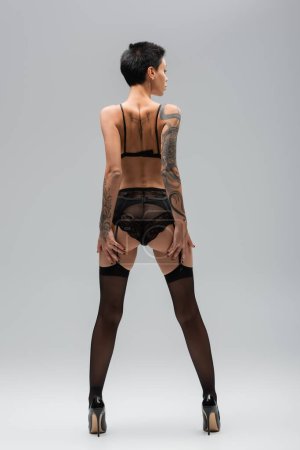 Rückansicht einer provokanten jungen Frau mit kurzen brünetten Haaren, sexy Gesäß und tätowiertem Körper im schwarzen BH, Spitzenhöschen, Strumpfgürtel, Strümpfen und High Heels auf grauem Hintergrund