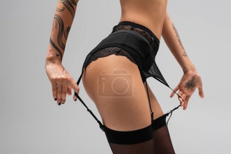 Foto de Vista recortada de mujer apasionada con sexy cuerpo tatuado tocando correas de liguero negro mientras posa en bragas de encaje y medias sobre fondo gris, fotografía erótica - Imagen libre de derechos