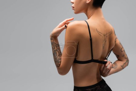back view of passionate young woman with sexy tattooed body unbuttoning black bra while posing on grey background, art of seduction, erotic photography, provoke 