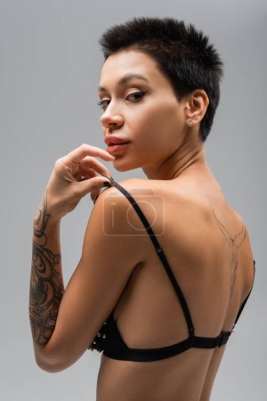 young and flirtatious woman with short brunette hair and sexy tattooed body touching strap of black bra while looking at camera on grey background, erotic photography