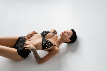 high angle view of sensual and sexy woman with closed eyes and tattooed body laying in bra with pearl beads and black lace panties on grey background, erotic photography