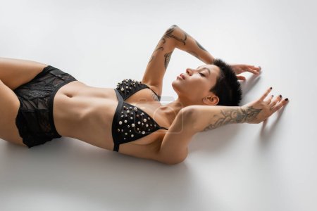 high angle view of seductive tattooed woman with short brunette hair and closed eyes laying in bra with pearl beads and lace panties on grey background, art of seduction