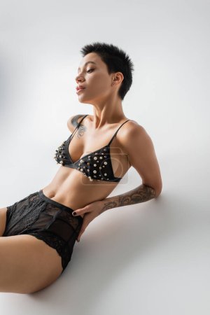 mujer joven y sexy con cuerpo tatuado y pelo corto morena sentado en sujetador con cuentas de perlas y bragas de encaje sobre fondo gris, fotografía erótica, arte de la seducción