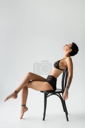 Foto de Vista lateral de mujer elegante con pelo corto morena y cuerpo tatuado sexy sentado en silla en sujetador negro con cuentas de perlas y bragas de encaje sobre fondo gris, fotografía erótica - Imagen libre de derechos
