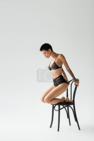 Photo for Full length of appealing young woman with short brunette hair and sexy tattooed body, wearing black bra with pearl beads and lace panties while posing on chair on grey background - Royalty Free Image