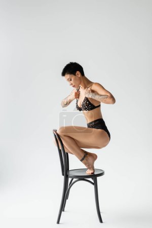 Photo for Full length of desirable woman with sexy tattooed body and short brunette hair, in bra with pearl beads and black lace panties posing on chair on grey background, art of seduction - Royalty Free Image