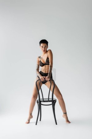 Photo for Full length of young seductive woman with short brunette hair and sexy tattooed body, in bra with pearl beads and lace panties looking at camera while posing with chair on grey background - Royalty Free Image