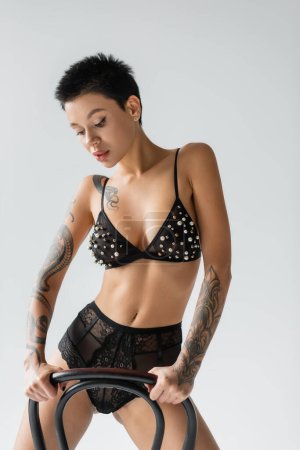 Photo for Young expressive woman with short brunette hair and sexy tattooed body posing near chair in black bra with pearl beads and lace panties on grey background, erotic photography - Royalty Free Image