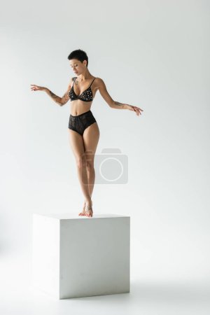Photo for Full length of appealing woman with sexy tattooed body and short brunette hair standing on white cube in black bra with pearl beads and lace panties on grey background - Royalty Free Image