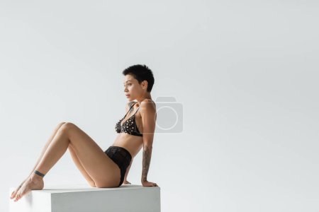 full length of young, sexy and tattooed woman with short brunette hair sitting on white cube in black bra with pearl beads and lace panties on grey background, erotic photography