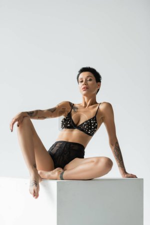 full length of stunning woman with sexy tattooed body and short brunette hair, wearing black bra with pearl beads and lace panties and looking at camera on white cube on grey background