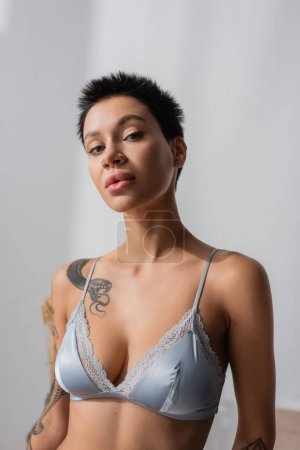 portrait of young ad sexy woman in silk bra, with tattooed body, short brunette hair and natural makeup looking at camera while posing in bedroom at home on blurred background