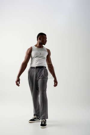 Volle Länge des trendigen jungen afroamerikanischen Mannes in ärmellosem T-Shirt und Hose, der vor grauem Hintergrund wegschaut, zeitgenössisches Shooting in lässiger Kleidung 