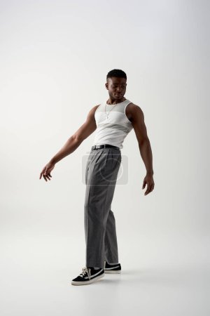 Longitud completa de hombre afroamericano joven de moda en pantalones, camiseta sin mangas y zapatillas de deporte de pie sobre fondo gris, sesión contemporánea con atuendo casual